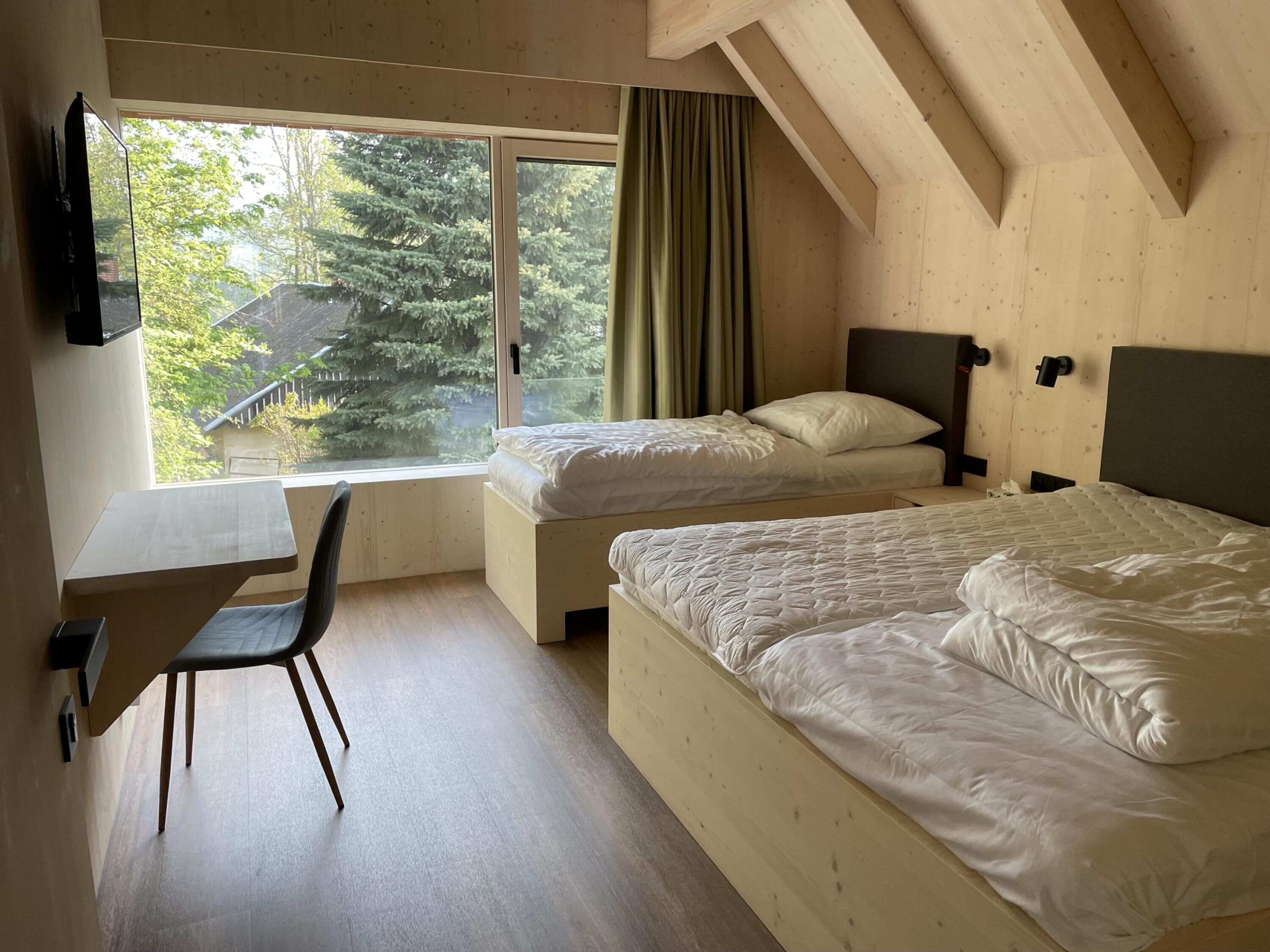 A room with two beds, a desk, and a chair. There is a window with a view of trees and a house.