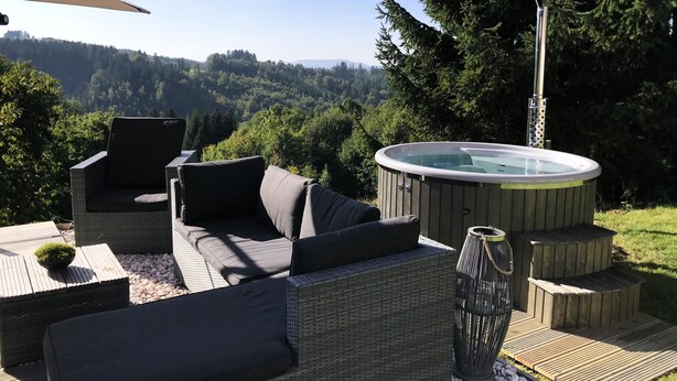 Un área de estar al aire libre con mobiliario de mimbre, un jacuzzi y vistas a un paisaje boscoso.