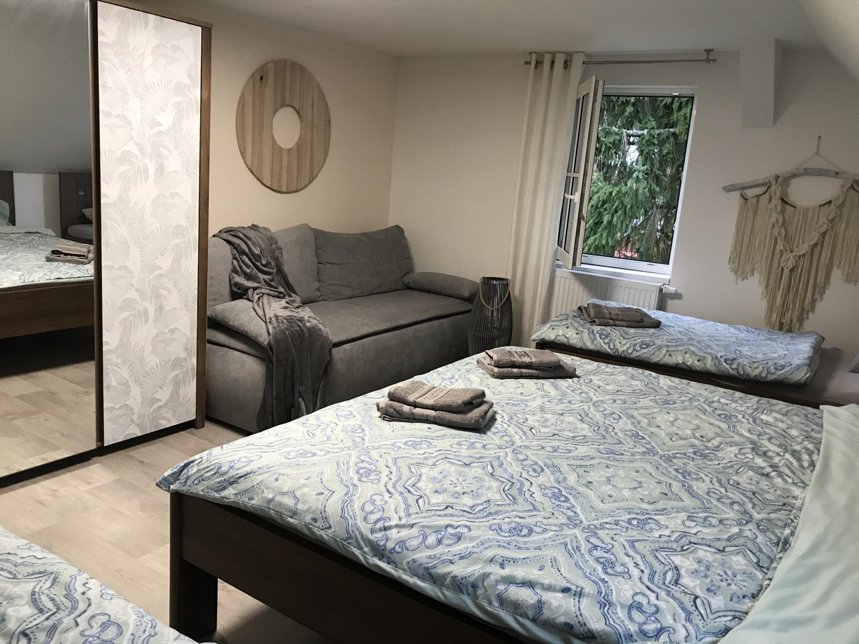 Quarto com espelho grande, sofá, duas camas com roupa de cama estampada, janela com cortinas e um enfeite de macramé na parede.