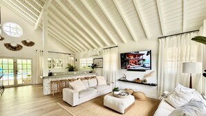 A spacious living room with a high ceiling, a large TV mounted on the wall, a cozy seating area with a sofa and ottoman, and a kitchen area with a bar counter. - WOW STYLISH! Next2Bahamar: Pickleball, PrvPool, HotTub + Beachlife & Restaurants (Nassau)