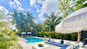 A poolside area with a white sofa set, a pool, and palm trees. - WOW STYLISH! Next2Bahamar: Pickleball, PrvPool, HotTub + Beachlife & Restaurants (Nassau)