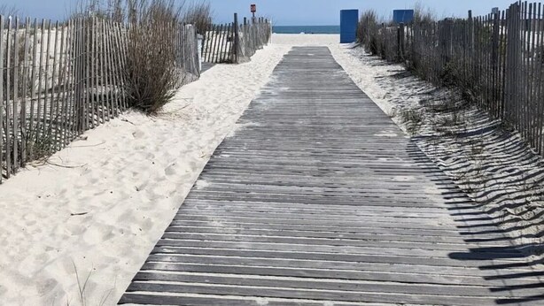 Una pasarela de madera que conduce a una playa con arena a ambos lados, una estructura azul en la distancia y un cielo azul despejado.