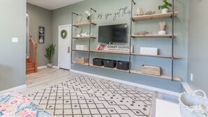 Una sala de estar con un televisor montado en la pared, estanterías de madera con objetos decorativos, un tapete estampado y un letrero que dice "Coast Guard Training Center Cape May".