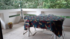 Un balcón con una mesa cubierta por un mantel floral, sillas blancas y una maceta con flores amarillas.