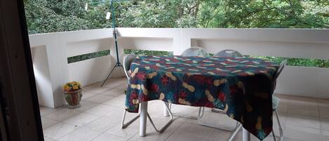A balcony with a table covered by a floral tablecloth, white chairs, and a potted plant with yellow flowers.