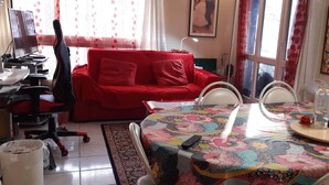 Una sala de estar con un sofá rojo, un sillón gamer, una mesa de comedor con mantel colorido y un televisor sobre un escritorio.