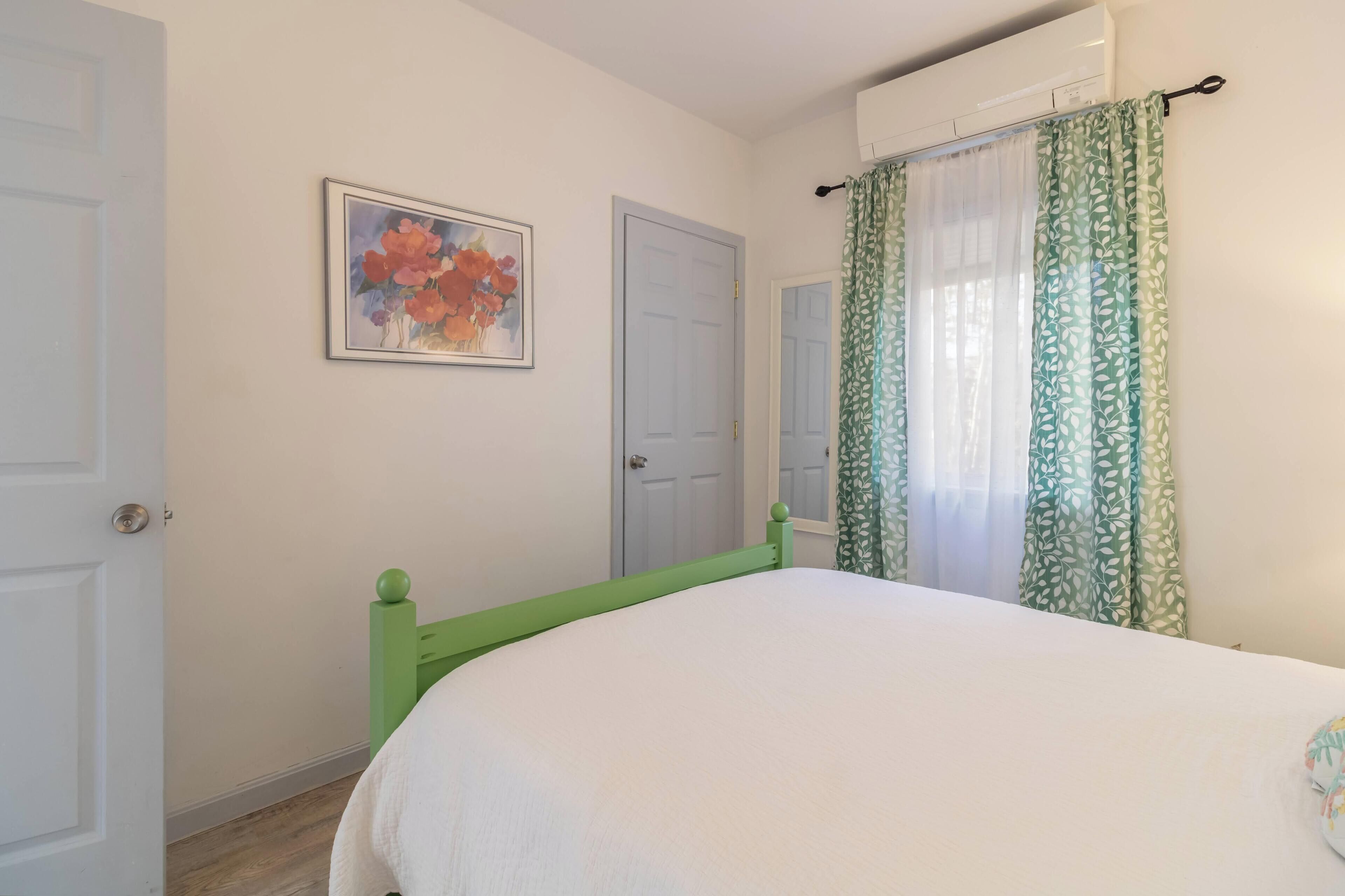 Una camera da letto con un letto, una porta, un condizionatore d'aria a parete e una finestra con tende a motivi verdi.