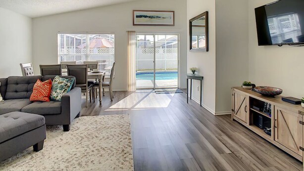 A modern living room with a gray sectional sofa, a dining area with a table and chairs, a TV mounted on the wall, and a view of a swimming pool through glass doors. - A Dark Siders dream in Vader's Vacation Villa.  (Davenport)