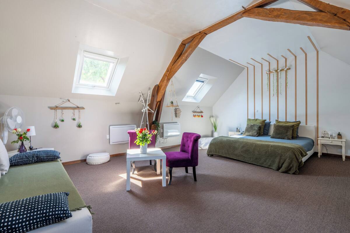 A bedroom with a sloped ceiling, a bed with a green blanket, a purple chair, a white table with a vase, and a window with a view of trees.