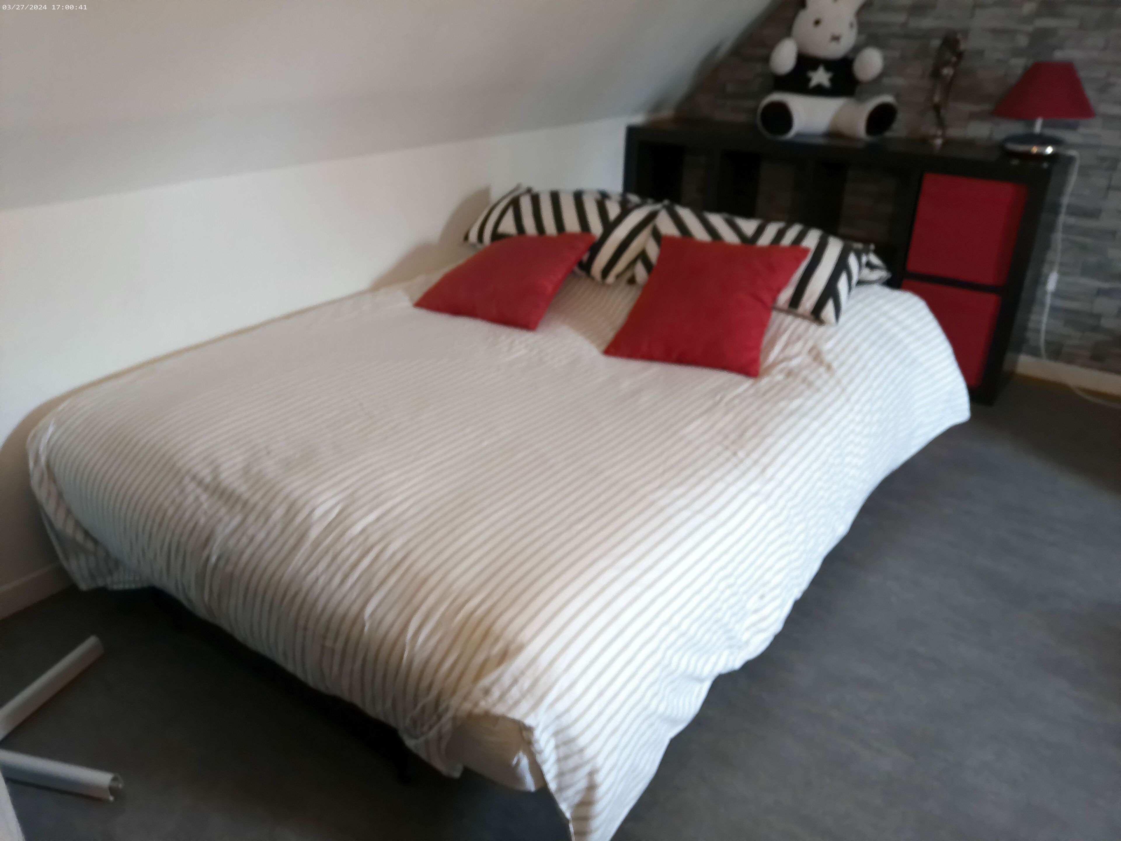 Een net opgemaakt bed met gestreepte lakens en rode kussens, een klein nachtkastje met een lamp, en een decoratief object aan de muur.