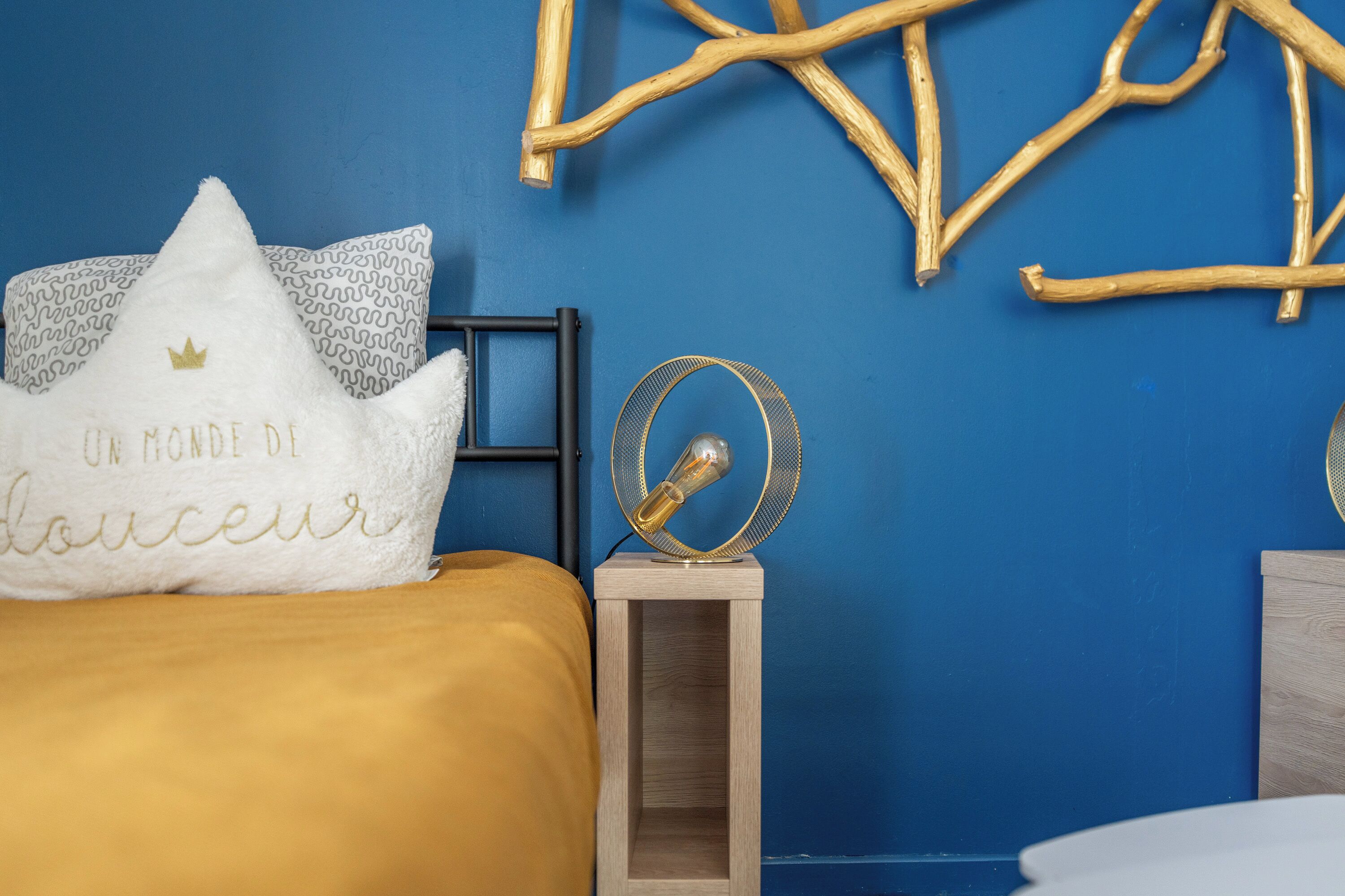 Una cama con una colcha dorada y un almohadón con texto. Hay una pequeña mesita de noche con una lámpara y un objeto decorativo encima. La pared está pintada de azul, y hay una rama de madera decorativa sobre la cama.