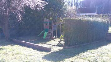 Ein Spielplatz mit Rutsche und Klettergerüsten, umgeben von einem Hecken und Bäumen.