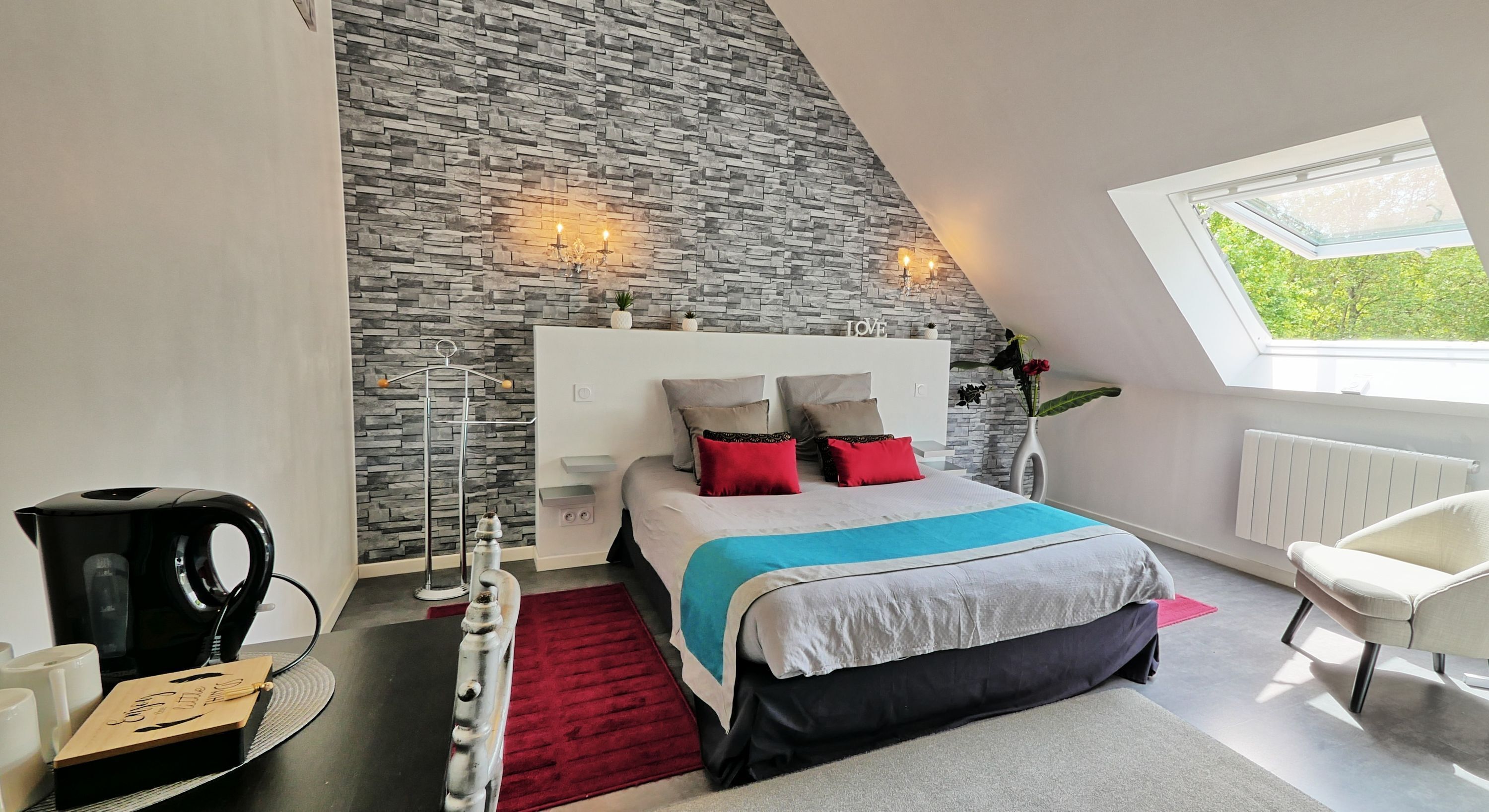 A modern bedroom with a stone wall, a bed with red and blue bedding, a white chair, and a skylight.