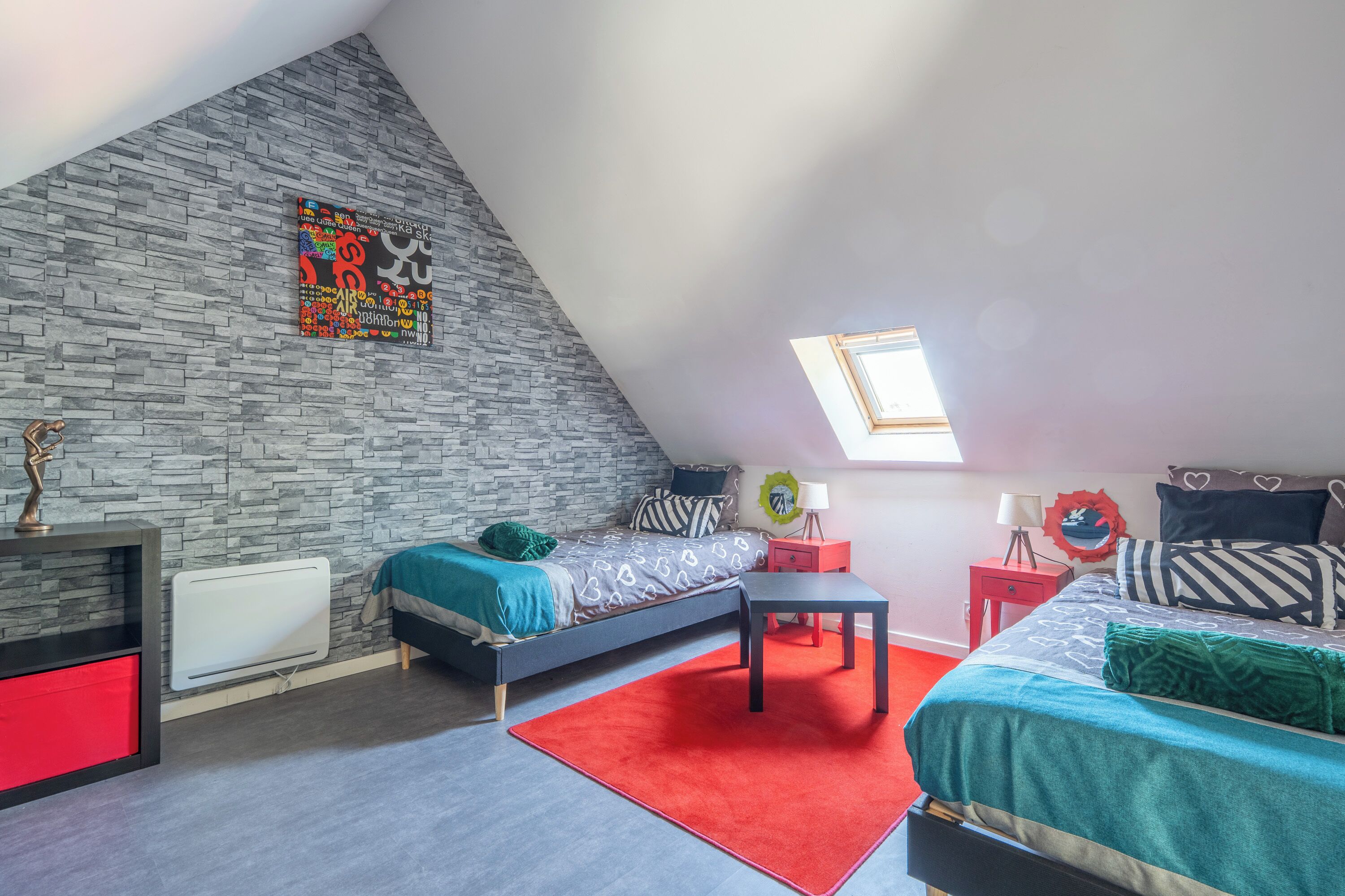 Une chambre à coucher aménagée dans un espace mansardé, avec un plafond en pente, un puits de lumière, un lit avec une literie à motifs, un tapis rouge et un mur en pierre orné d’une œuvre d’art colorée.