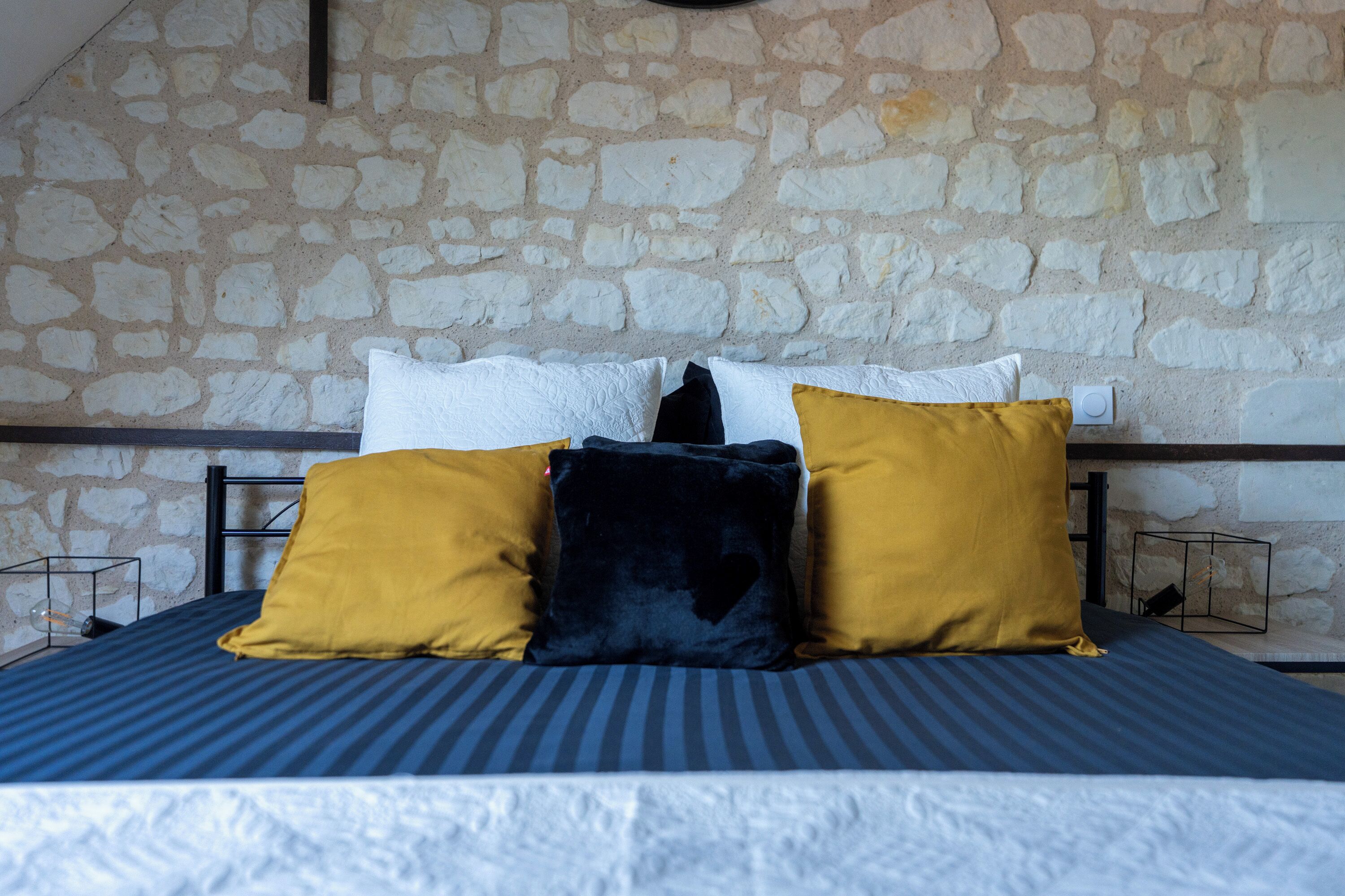 A bed with a striped blue and white bedspread, three pillows in yellow, black, and white, and a stone wall behind.