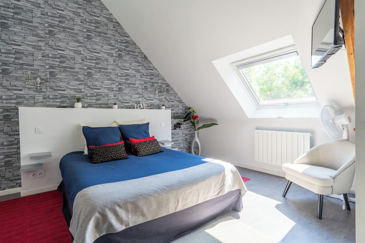 A bedroom with a sloped ceiling, a skylight, a bed with blue bedding, a white chair, and a radiator.
