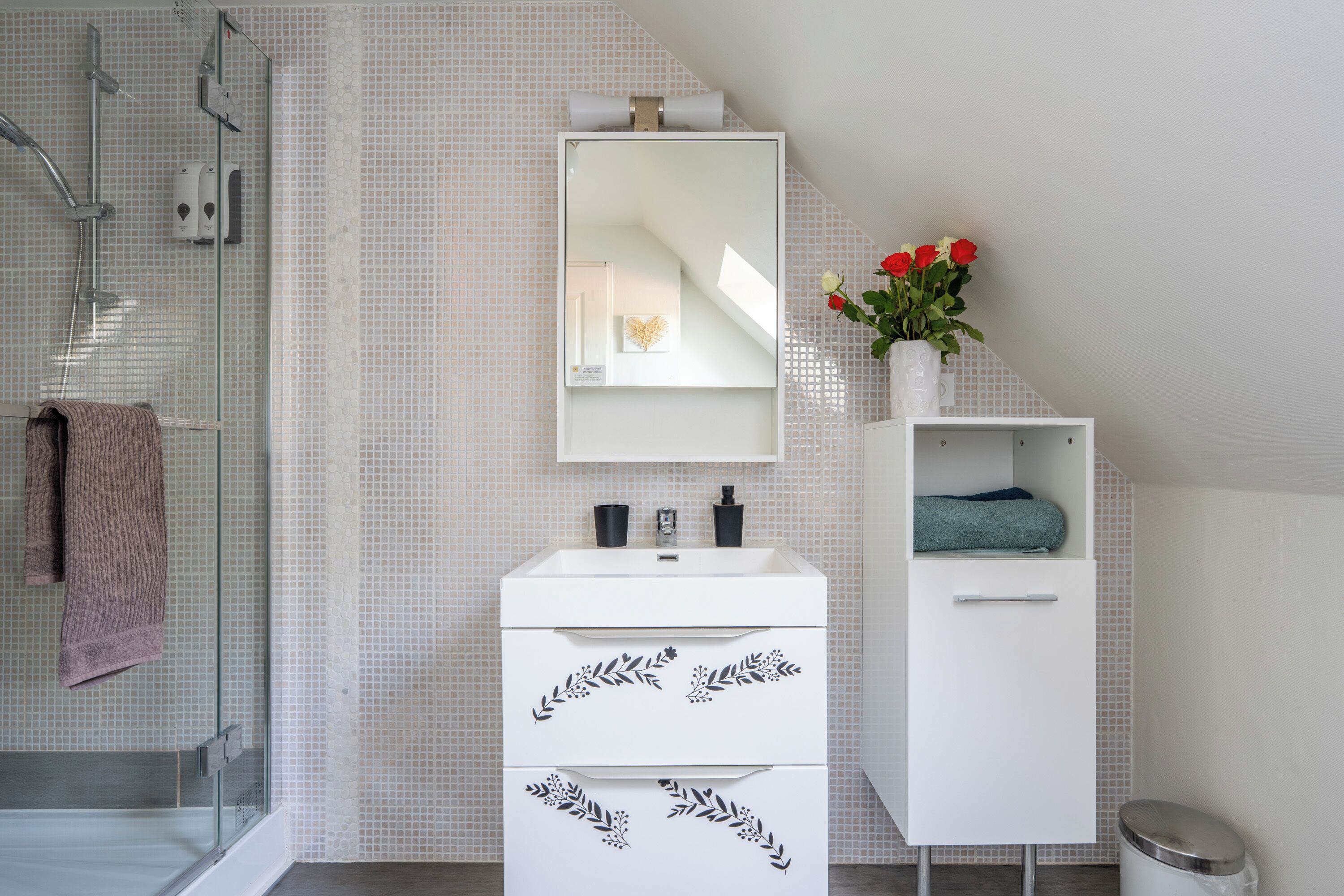 Un baño moderno con cabina de ducha de vidrio, un tocador blanco con espejo y un pequeño estante con un florero.