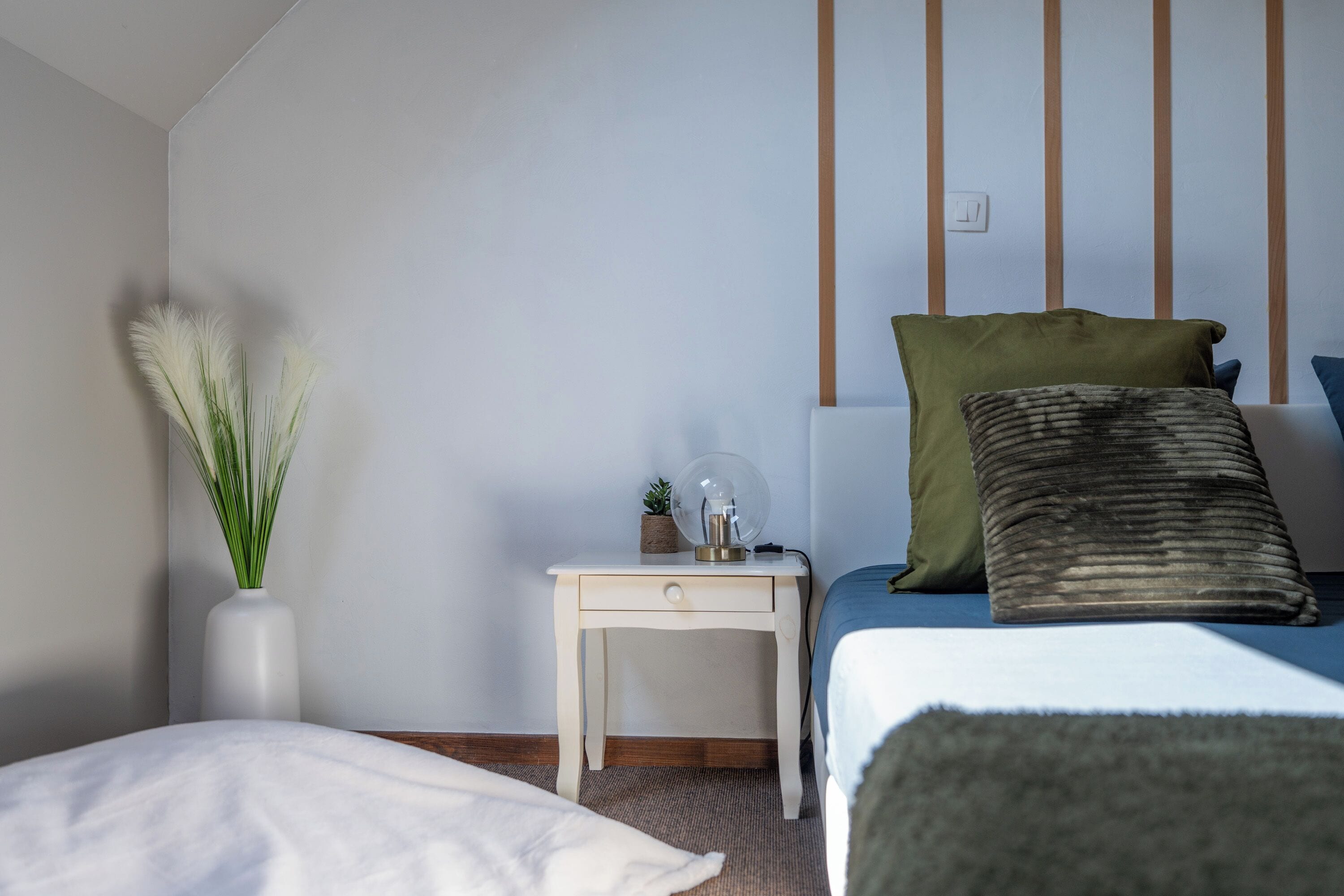 Een slaapkamer met een bed, nachtkastje, lamp en een vaas met planten.