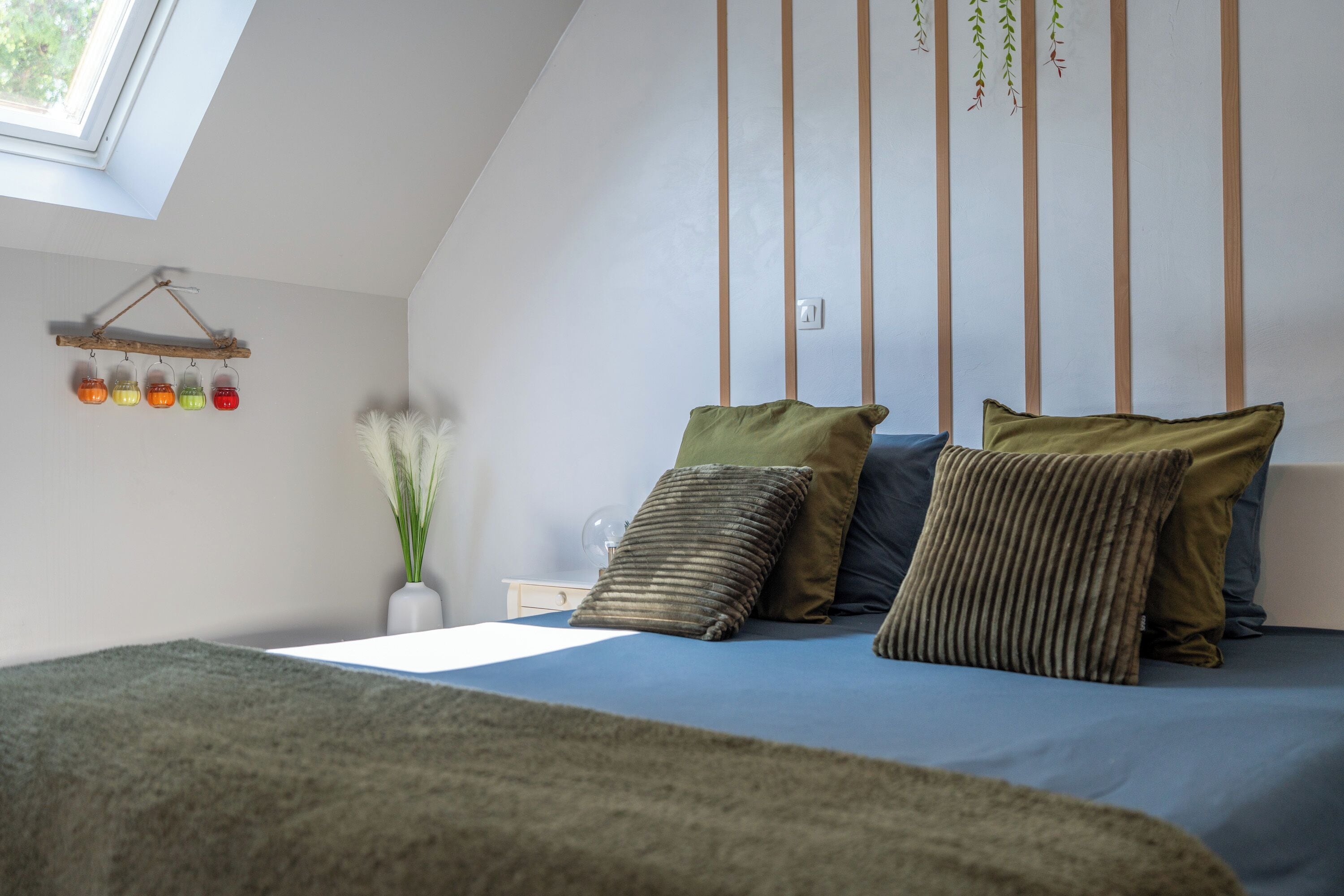 A bedroom with a sloped ceiling, a bed with multiple pillows, a bedside table with a lamp, and a wall-mounted shelf with jars.