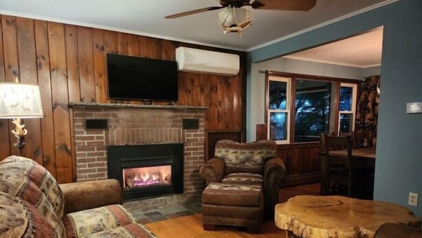 A cozy living room with a fireplace, wooden walls, a ceiling fan, and a view of the outdoors. - West Mountain Ski & Adventure Home w/ Hot Tub
 (Queensbury)