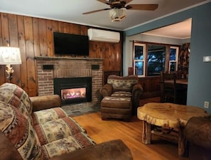 A cozy living room with a fireplace, wooden walls, a ceiling fan, and a view of the outdoors.