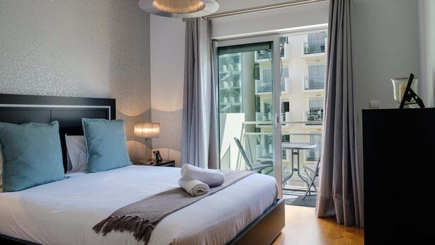 A modern hotel room with a large bed, a nightstand, a lamp, and a view of the outside balcony.