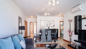 A modern living room with a sofa, coffee table, and a TV. There is a dining area with chairs and a table, and a kitchen in the background.