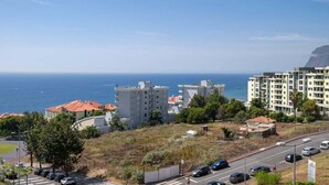 A coastal residential area with modern buildings, a road with parked cars, and a view of the sea.