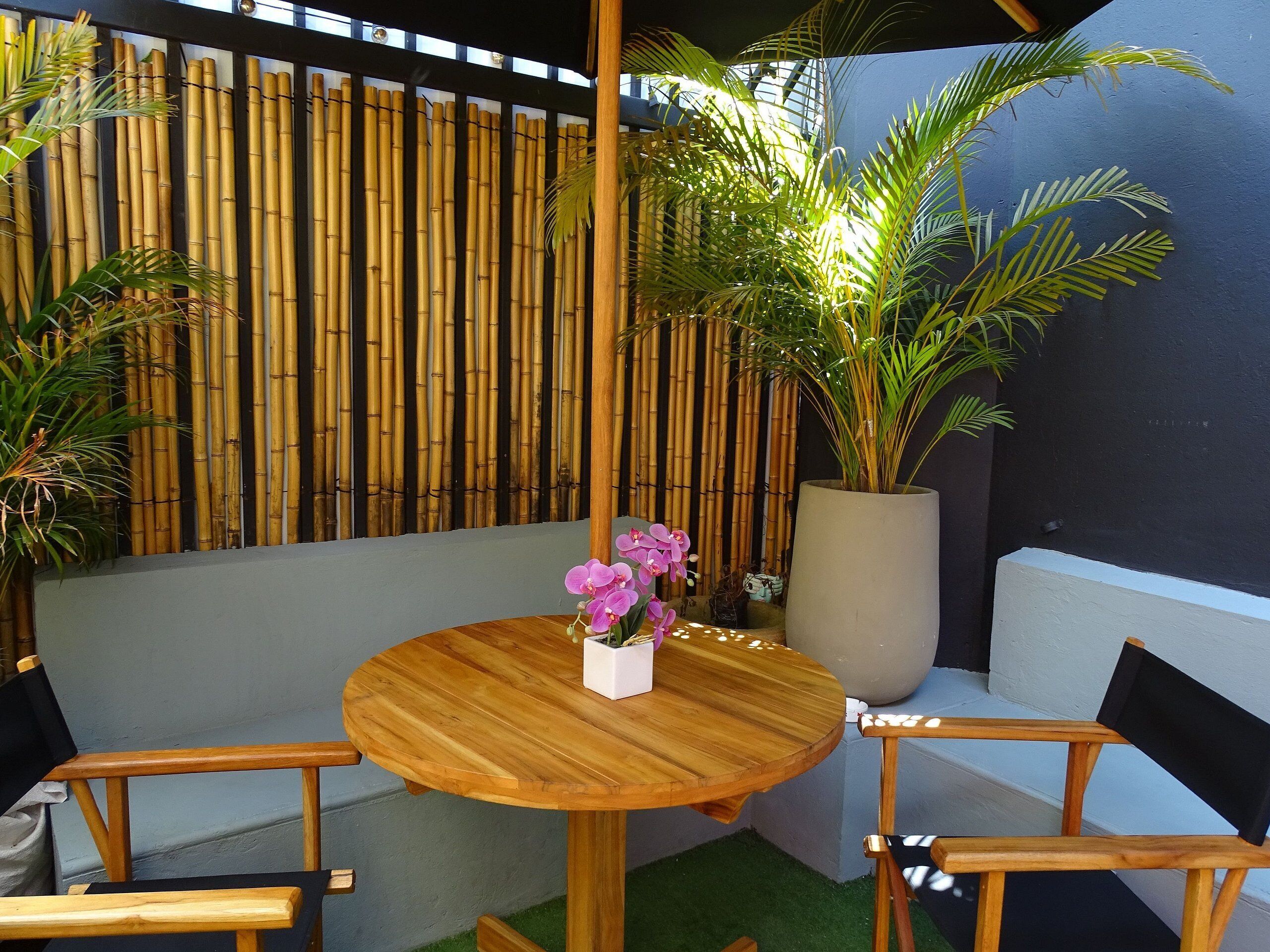 Une table en bois avec des chaises, une plante en pot et une clôture en bambou dans une cour.