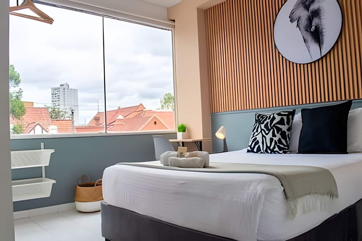 A bedroom with a bed, bedside table, lamp, and a view of buildings through a large window.