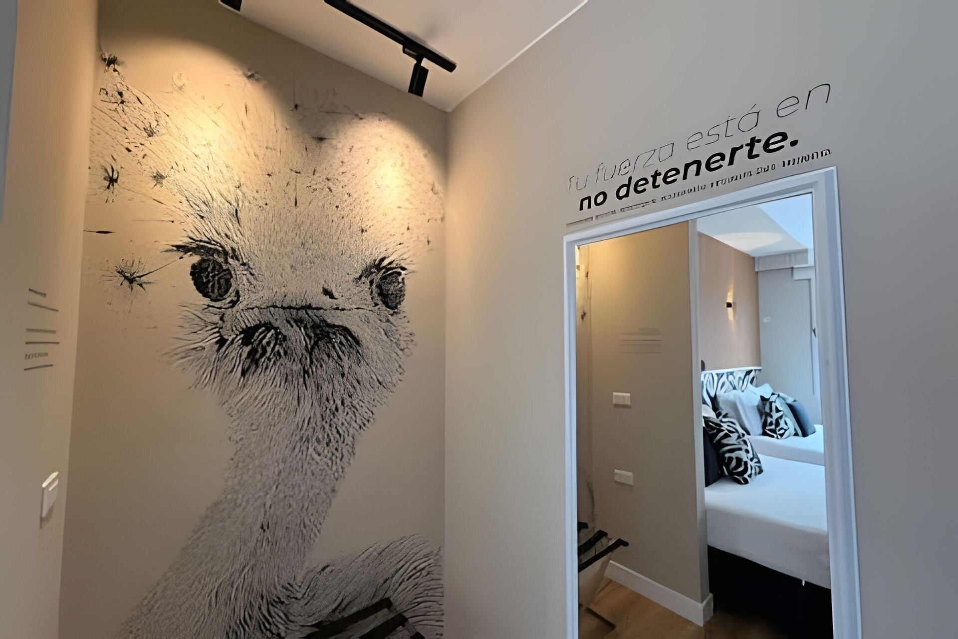 Una pared con una gran obra de arte texturizada del rostro de un animal, un vano que da a una habitación con una cama y almohadones estampados, y una pared con texto.