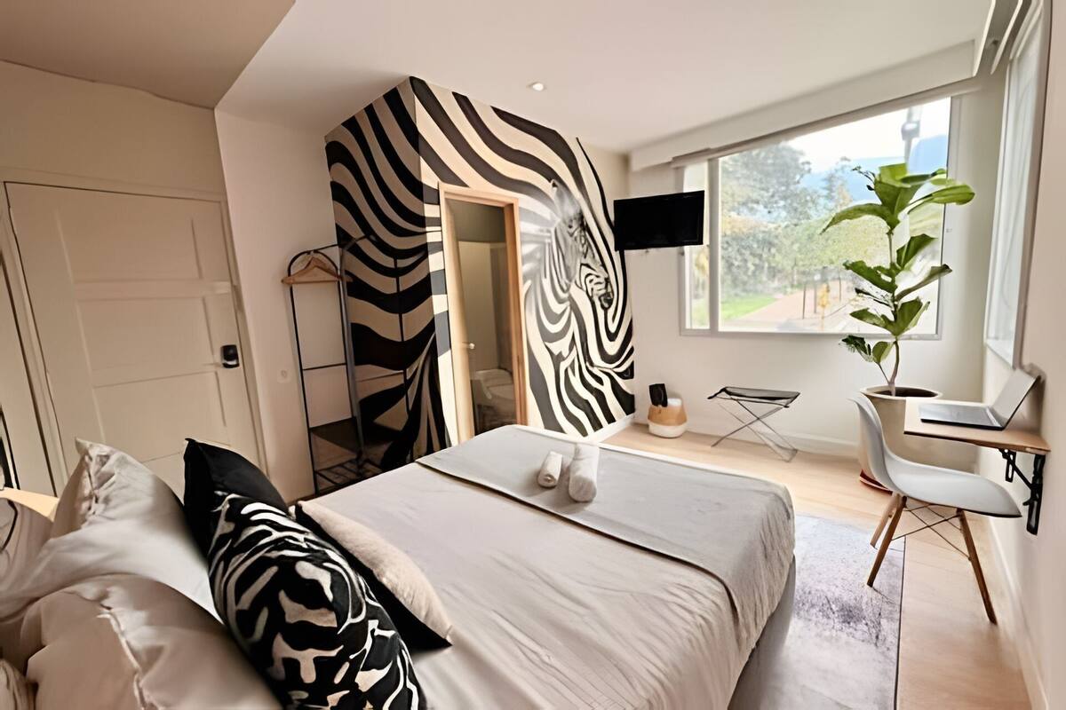 A modern bedroom with a zebra-striped wall, a bed with matching pillows, a desk with a laptop, and a large window.