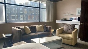 A modern hotel room with a large window, a sofa, a chair, a small table, and a kitchenette area.