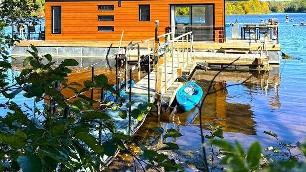 A modern houseboat with a wooden exterior, a glass balcony, and a dock extending into a lake.