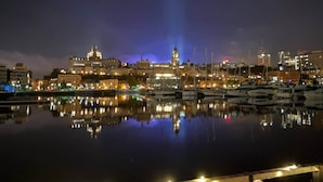 A nighttime cityscape with a marina and boats, a life preserver on a railing, and string lights.