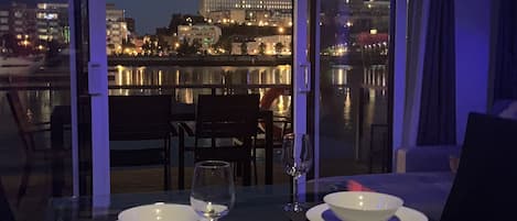 Una mesa de comedor con dos cuencos y copas de vino, con vistas a un paisaje urbano con edificios iluminados de noche.