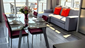 A modern room with a dining table set for two, a sofa with orange cushions, and a view of a waterfront.