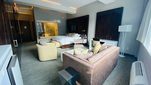 Une chambre d’hôtel moderne avec un grand lit, un canapé, une table basse et un téléviseur. Il y a une grande fenêtre et une porte qui donne sur une autre pièce.