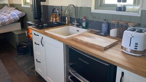 A compact kitchen with a stove, sink, and toaster. There is a window above the sink, and a bed with a plaid blanket is visible in the background.
