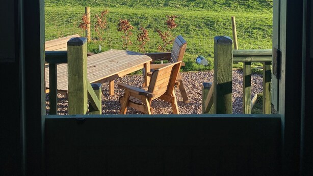 A view from a room with a wooden deck and a table, overlooking a green field and a clear blue sky.