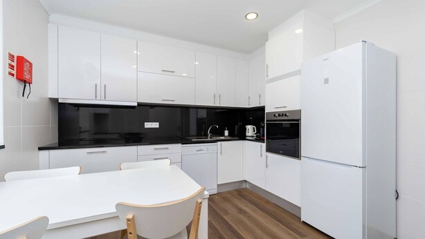 Eine moderne Küche mit weißen Schränken, einer schwarzen Arbeitsplatte, einem weißen Esstisch mit Stühlen sowie einem eingebauten Backofen und Kühlschrank.
