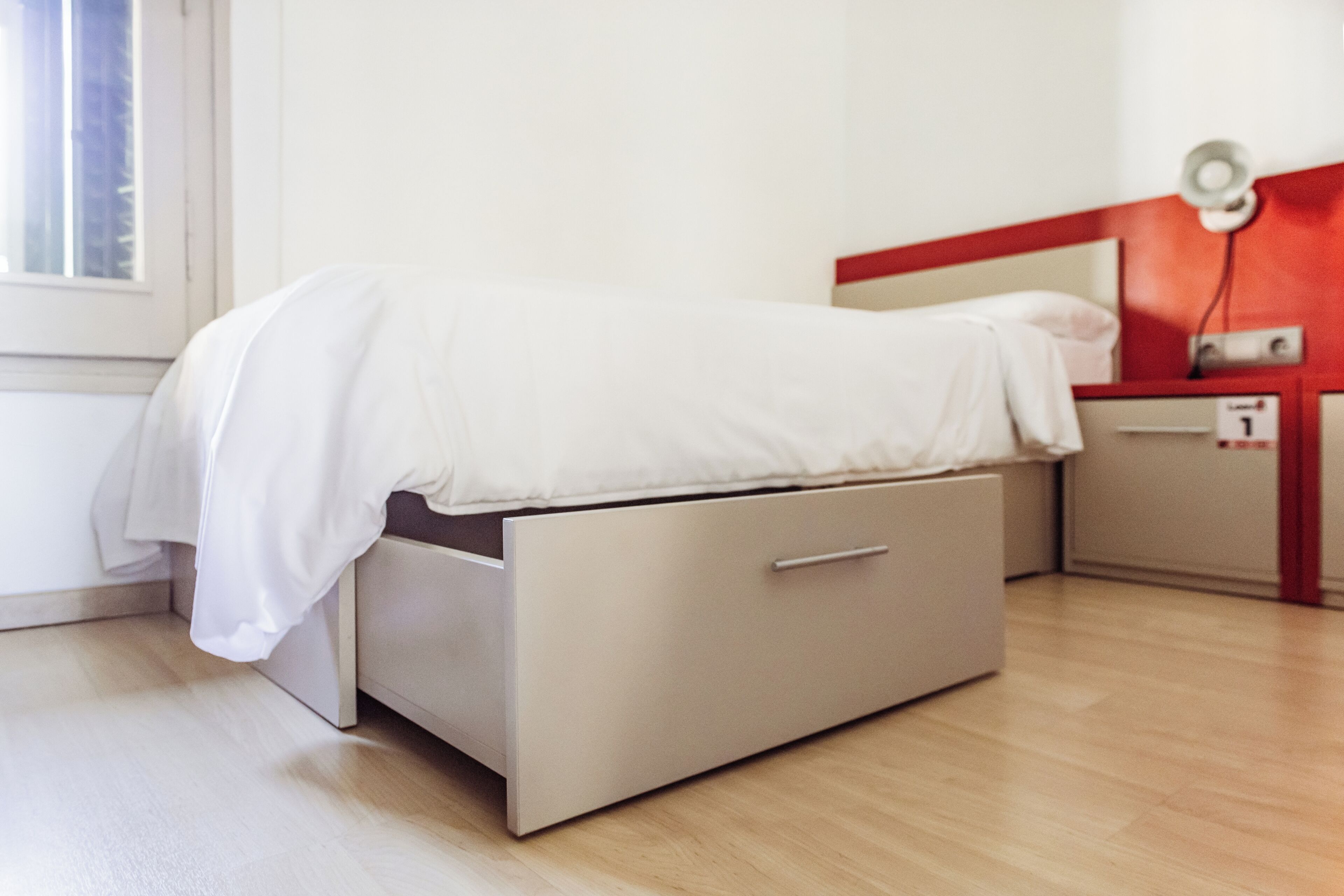 Una cama individual con un cajón para guardar objetos debajo, una mesita de noche con lámpara y una ventana con persianas.