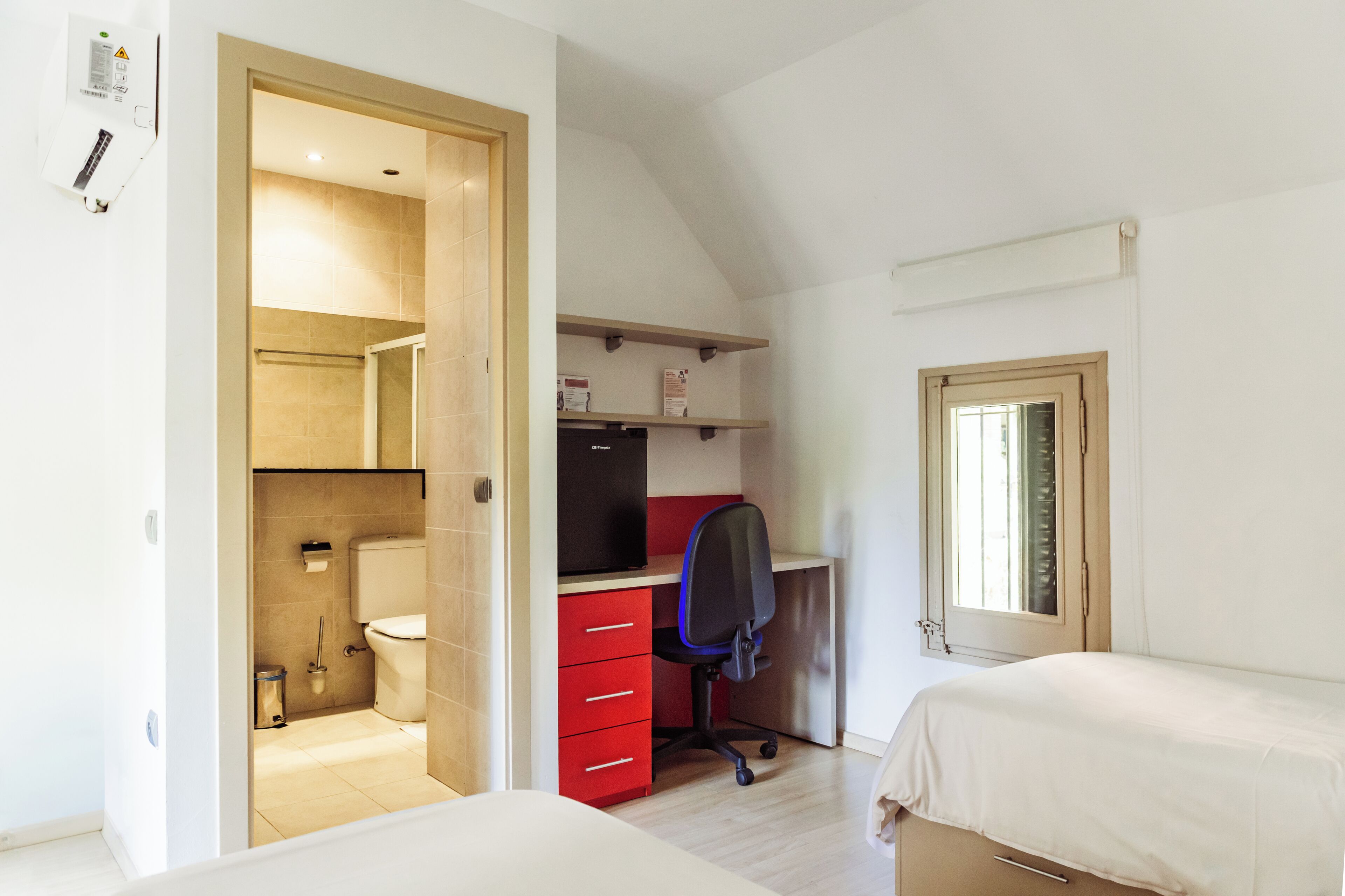 Una camera da letto con un letto, una scrivania con sedia, un bagno piccolo con il water e una finestra.