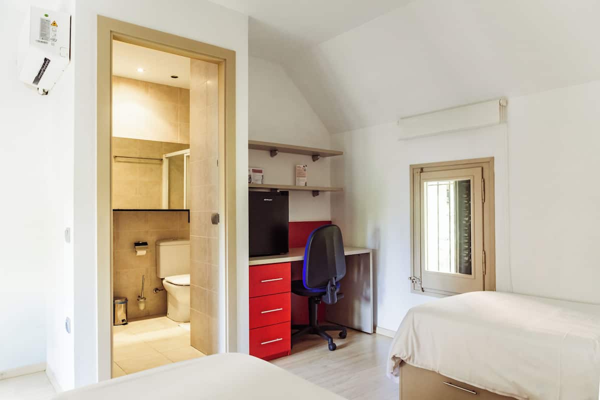 Un dormitorio con cama, un escritorio con silla, un baño pequeño con inodoro y una ventana.