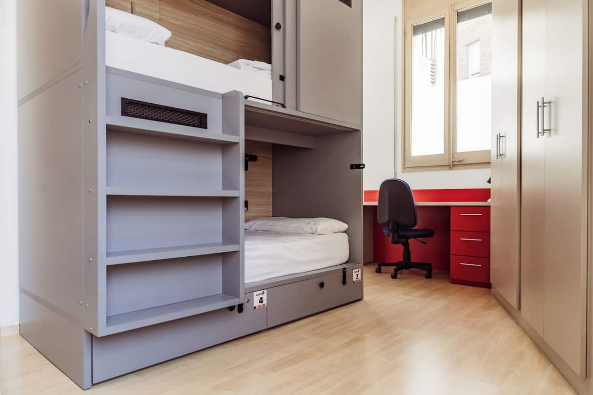 Una cama litera con cajones para guardar cosas, un escritorio y una silla en una habitación con suelo de madera y una ventana.