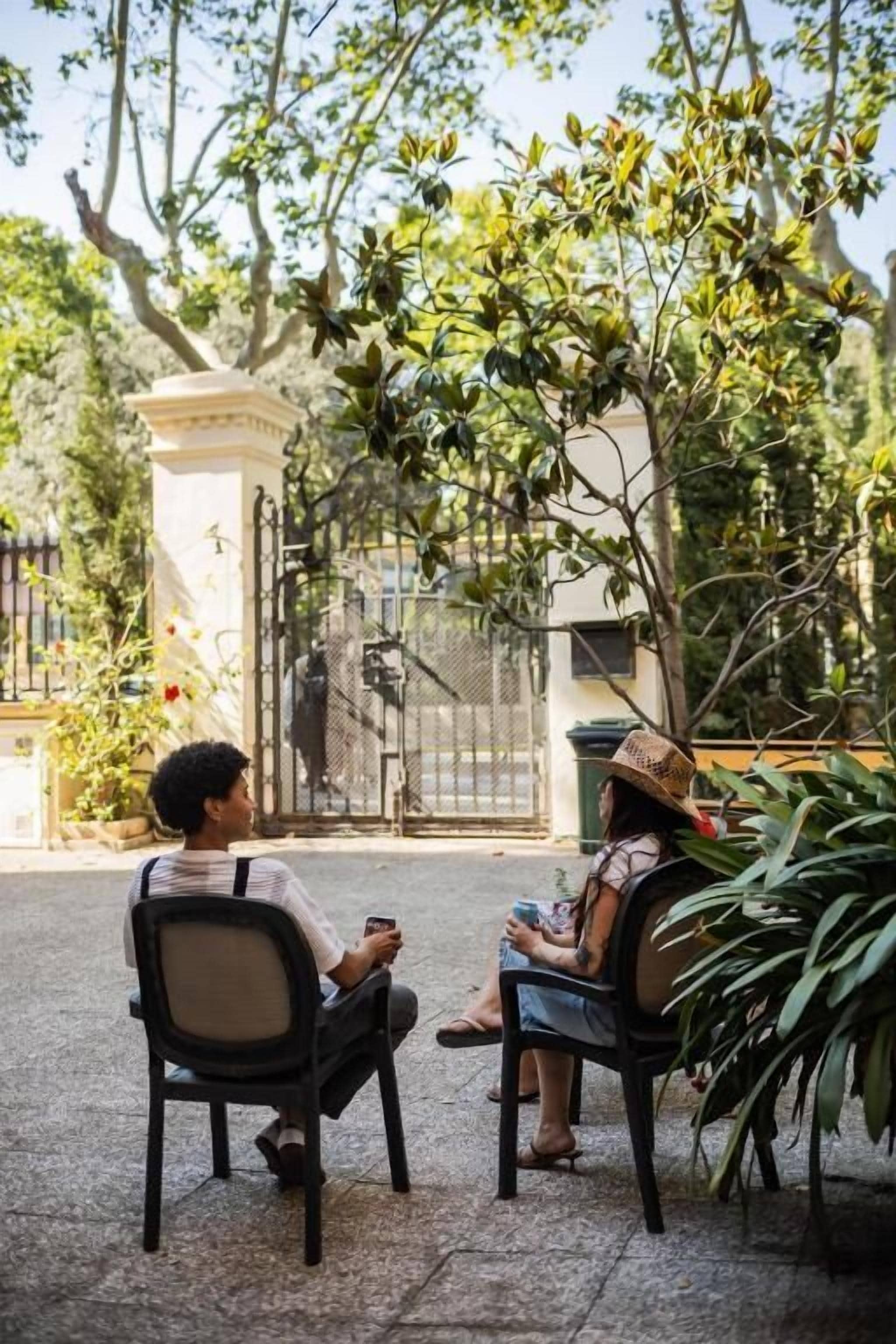 Two people seated on outdoor chairs, a gate, and surrounding greenery.