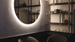 Une pièce faiblement éclairée, avec un luminaire circulaire au plafond, des étagères remplies de théières et une petite table entourée de chaises.
