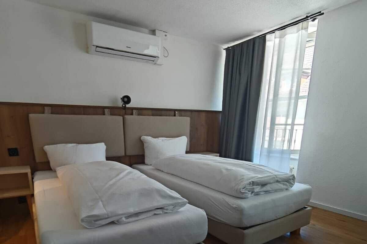 Ein Hotelzimmer mit zwei Betten, Holzboden, einem Vorhangfenster und einer Klimaanlage an der Wand.