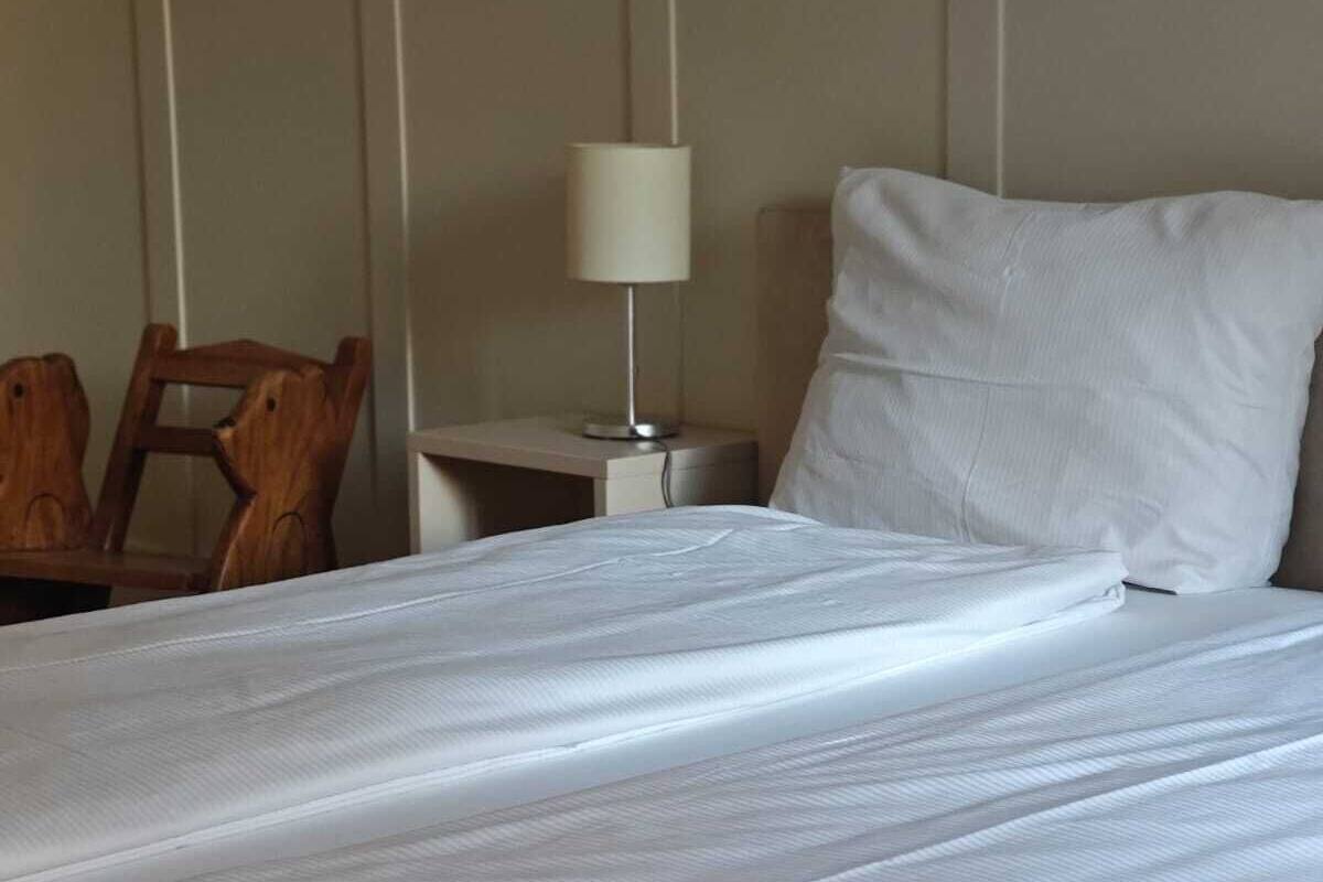 Ein ordentlich bezogenes Bett mit weißen Leinen, ein Holzbstuhl, ein Nachttisch mit Lampe und eine schlichte Wand.