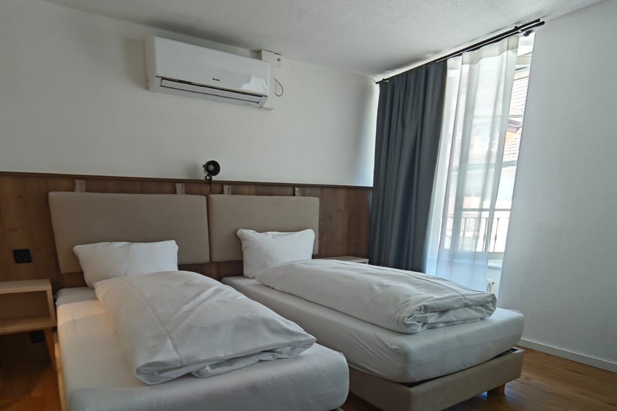 Een hotelkamer met twee bedden, een houten vloer, een raam met gordijnen en een airconditioningsunit aan de muur.